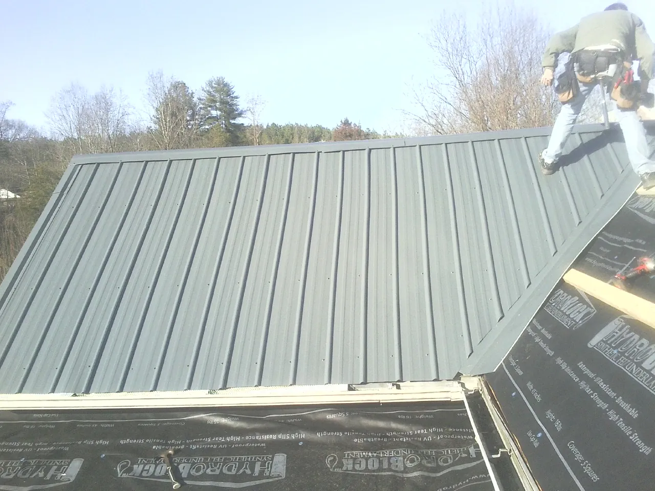 Crew installing dark green standing seam panels over synthetic underlayment in West Dennis