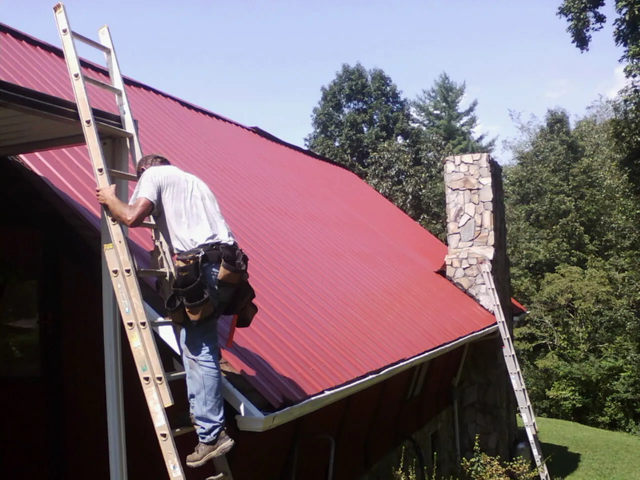 Roofer on a ladder beside a red metal roof and stone chimney — Soffit & Fascia Repair by Roofing Brothers in West Dennis